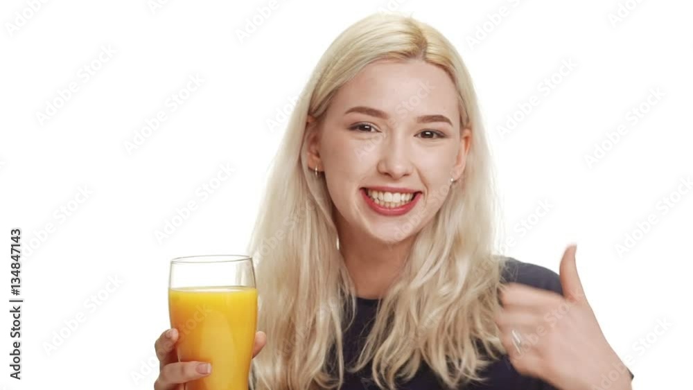 Young adorable blonde Caucasian teenage girl holding glass with orange juice smiling winking showing ok with thumb on white background in slowmtoion Young adorable blonde Caucasian teenage girl holding glass with orange juice smiling winking showing ok with thumb on white background in slowmtoion