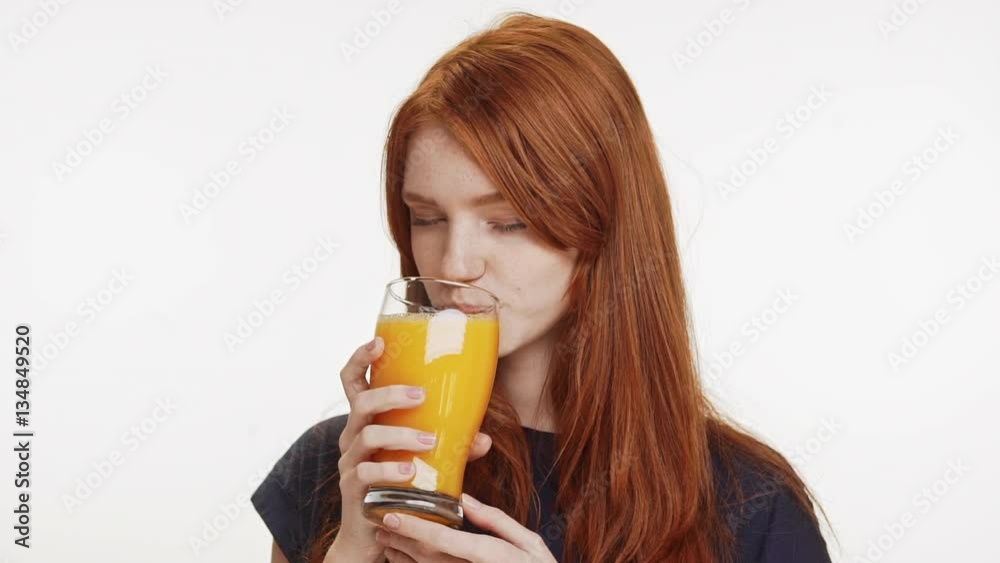 Young smiling cute foxy girl wearing dark blue shirt drinking orange juice from glass on white background in slowmotion Young smiling cute foxy girl wearing dark blue shirt drinking orange juice from glass on white background in slowmotion