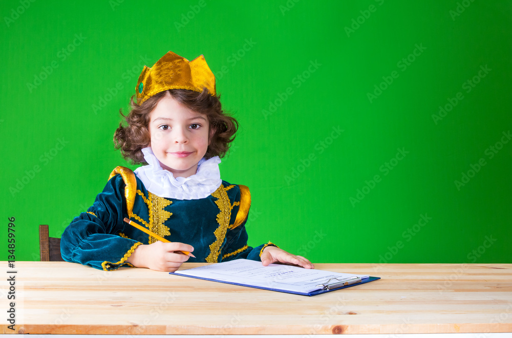 Little cute king sits at a table and writes in a notebook. Close-up. Green background. Little cute king sits at a table and writes in a notebook. Close-up. Green background.