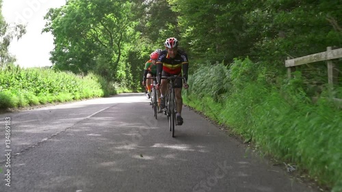 A stabilised view of a group of cycling athletes out on a training ride on country roads in the UK countryside on a sunny day. 