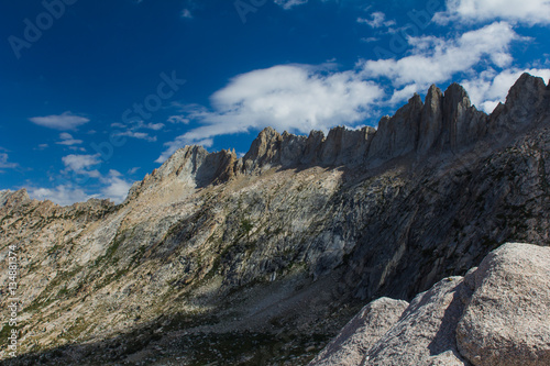 Blue sky and fluffy clouds hang above jagged granite peaks over a deep forested valley in California's high sierra