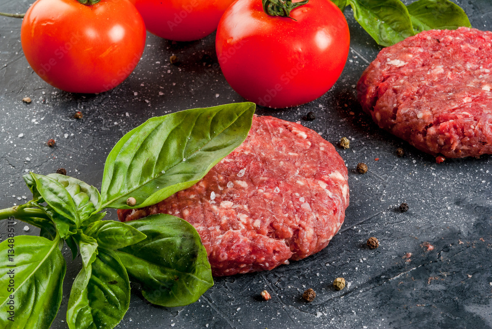 Fresh raw home-made minced beef steak burger with spices, tomatoes and basil, on a stone table, copy space, top view