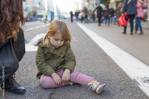 three years old blonde lovely caucasian girl child, with crying and sad expression and tear in face, next to mother, sitting on grey asphalt at pedestrian Gran Via Street in Madrid city, Spain
