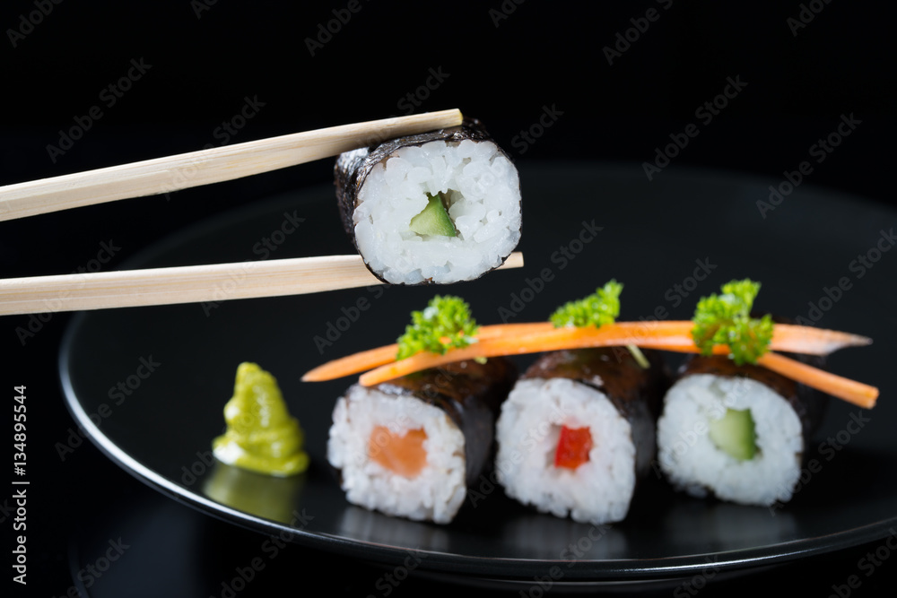 Sticks holding roll with cucumber on the background of fresh rolls with salmon, cucumber and pepper on a plate, served with carrot slices and parsley, black background Sticks holding roll with cucumber on the background of fresh rolls with salmon, cucumber and pepper on a plate, served with carrot slices and parsley, black background