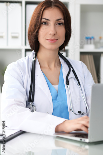 Happy brunette female doctor sitting at the table and filling up medical history form. Medicine, healthcare and help concept. Physician ready to examine patient