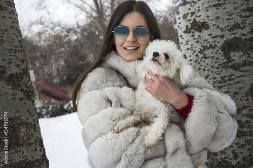 Beautiful winter close-up of a young girl in the winter snowy scenery. Woman wearing a white fur coat. Woman holding her dog. Wearing winter sunglasses.