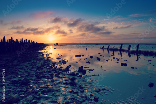 Beautiful sunny red colorful sunset on the lake with stones, wooden posts and reflection, natural seasonal summer vacation background