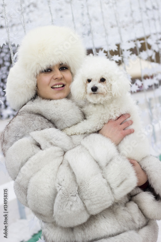 Winter beauty. Smiling young woman in the wintertime with her dog. Woman wearing white winter fur coat while holding her dog. Christmas decorations in the background.