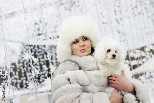 Winter beauty. Close up of a smiling young woman in the wintertime with her dog. Woman wearing white winter fur coat while holding her dog. Christmas decorations in the background.