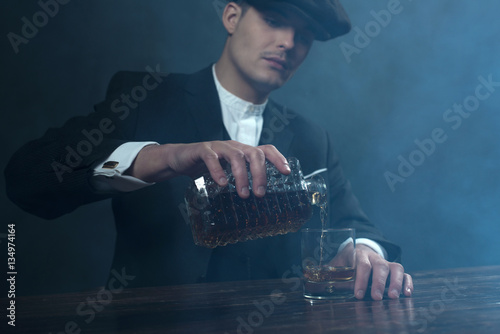 Retro 1920s english gangster with flat cap pouring whiskey. Peak