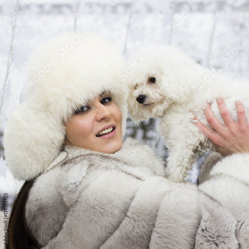 Winter beauty. Close-up of a young woman in the wintertime with her dog. Woman wearing white winter fur coat while holding her dog. Christmas decorations in the background.
