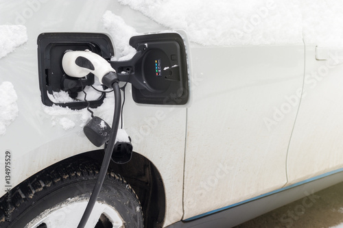 Electric car charging in winter time. Snow covered electric car with plugged electricity cabel