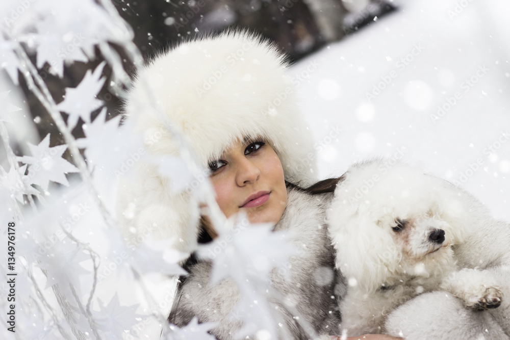 It is snowing! Beautiful woman in the winter time with her dog. Woman wearing a winter coat and a fur hat. Snowfall around her. It is snowing! Beautiful woman in the winter time with her dog. Woman wearing a winter coat and a fur hat. Snowfall around her.
