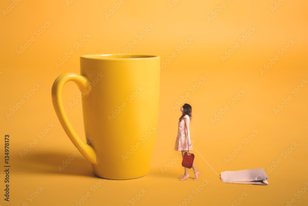Surrealism design. Girl holding a tea bag in his hand. Miniature people. Yellow empty cup on a yellow background. Young girl makes tea. - obrazy, fototapety, plakaty 