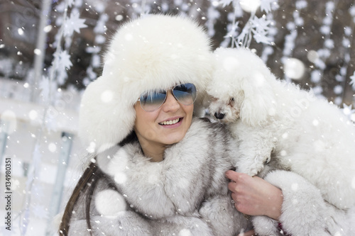 Beautiful woman playing with her dog outdoors. Winter time. Woman wearing a white fur coat. Christmas decorations in the background. Snowfall.