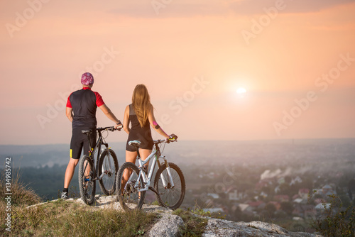 Fototapeta Naklejka Na Ścianę i Meble -  Rear view couple with sports bicycles standing on the top of a hill enjoying the sunset. Blurred background with copy space