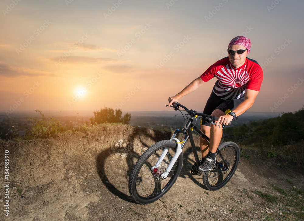 Guy in glasses riding on the mountain bicycle on a trail against evening sky with bright sun on the sunset