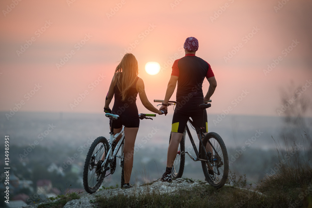 Naklejka premium Rear view cyclist couple with mountain bikes standing on a rock holding hands enjoying the sunset. Bright sun in between. Blurred background