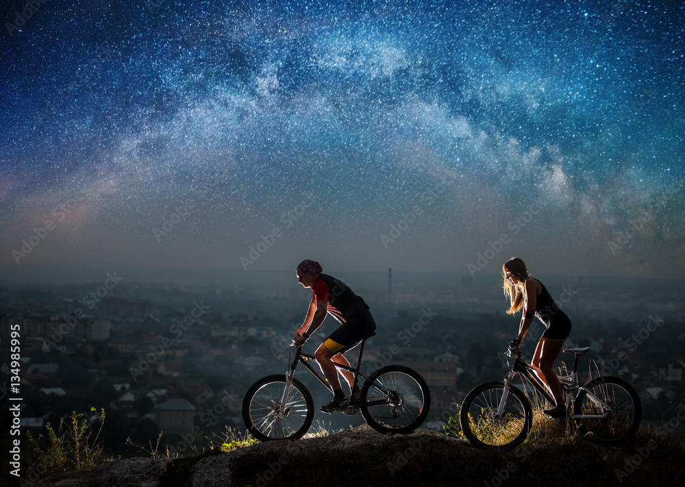 Fototapeta premium Sporty friends riding a bicycles on the mountain hill at night under starry sky and Milky way. Below is a city in the distance