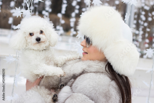 Beautiful woman playing with her dog outdoors. Winter time. Woman wearing a white fur coat. Christmas decorations in the background.
