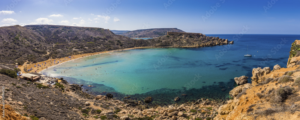 Malta - Panoramic view of Ghajn Tuffieha sandy beach with sail boat, blue sky and crystal clear green sea water 
