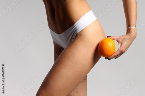Mid section of woman with sport body holding orange near her leg, anti-cellulite concept 