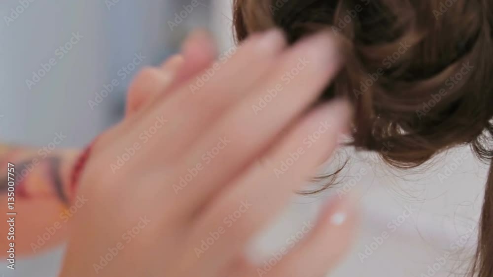Close up shot. Top knot hair design - hair stylist in glasses finishing hairstyle for young pretty woman and using bobby pins for fixing hair in white make up room. Beauty and haircare concept