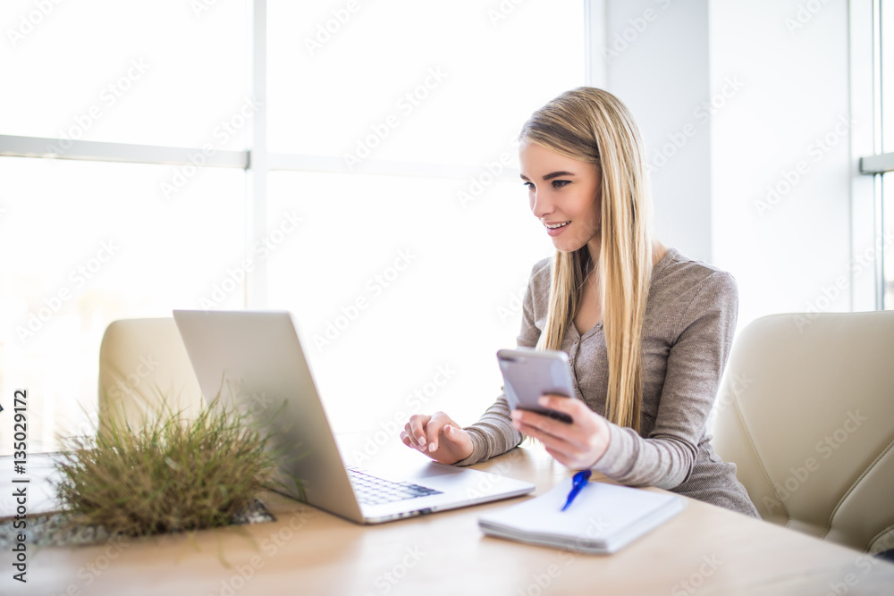 Portrait of a young business woman with phone in hand using laptop at office Portrait of a young business woman with phone in hand using laptop at office