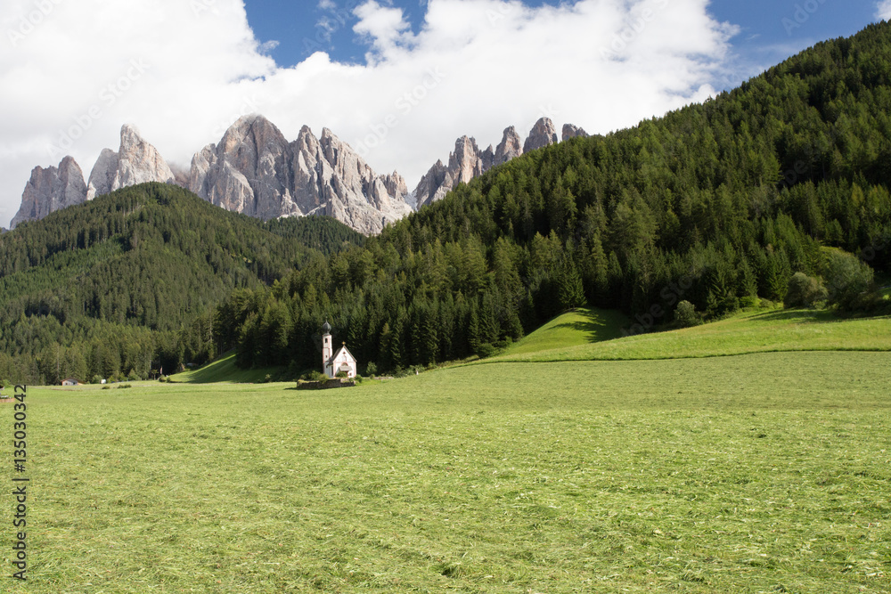 The symbol of the valley Val di Funes - church of Santa Maddalena. Rocky peaks and forested mountains surrounded by green Alpine meadows. Tirol, Dolomites. Sunny warm autumn day