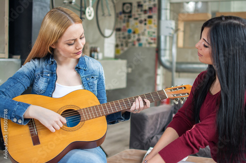 Two young female friends. One girl playing guitar and the other one listening.
