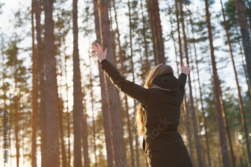 teen girl raised hands from behind in winter pine forest in sunset, freedom or victory concept