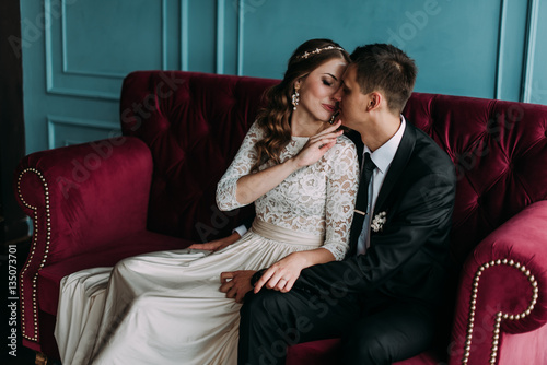 cute wedding couple in the interior of a classic studio posing at the sofa . hey kiss and hug each other, holding hands looking at each other