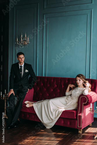 cute wedding couple in the interior of a classic studio posing at the sofa . hey kiss and hug each other, holding hands looking at each other © dashamuller