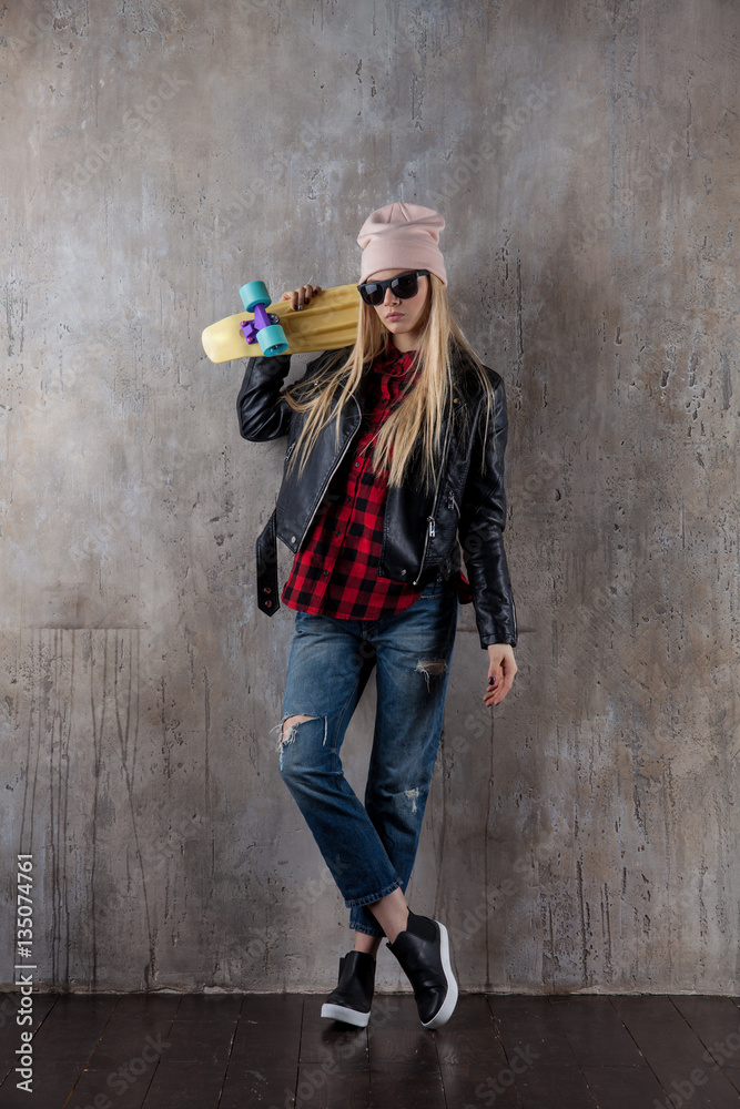 Blond teenager girl in black sunglasses and pink hat with blue skateboard. In front of concrete wall.