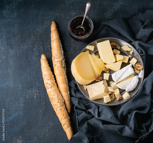 Cheese plate. Assortment variety of cheese with walnuts, jam and bread on vintage metal plate with textile over dark blue canvas as background. Top view with space. Appetizer theme