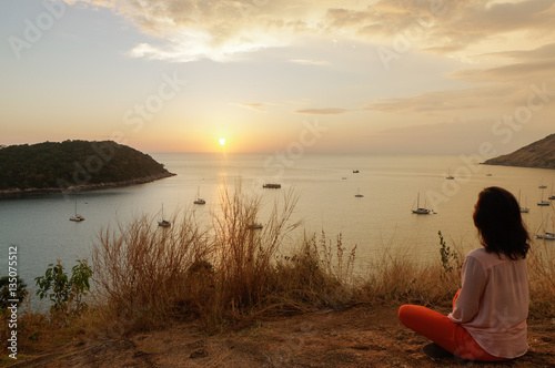Young girl sitting in yoga lotus meditation position front to seaside on the rocks an watching the sun goes down in a golden hour. sunset over sea.