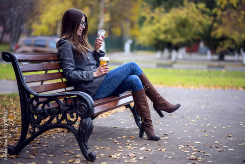 Girl reads sms, rewritten smartphone outdoors in spring or autumn, on the bench, with coffee tea, breakfast, concept, glasses, urban fashion, lifestyle, brunette.