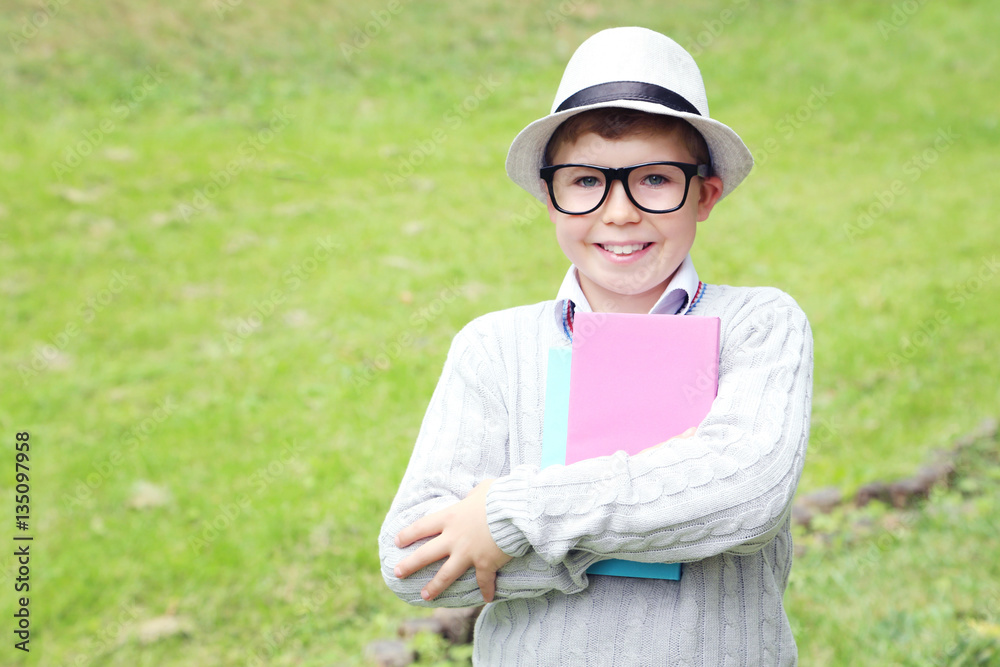 Fototapeta premium Portrait of young boy with eyeglasses, books and hat in the park