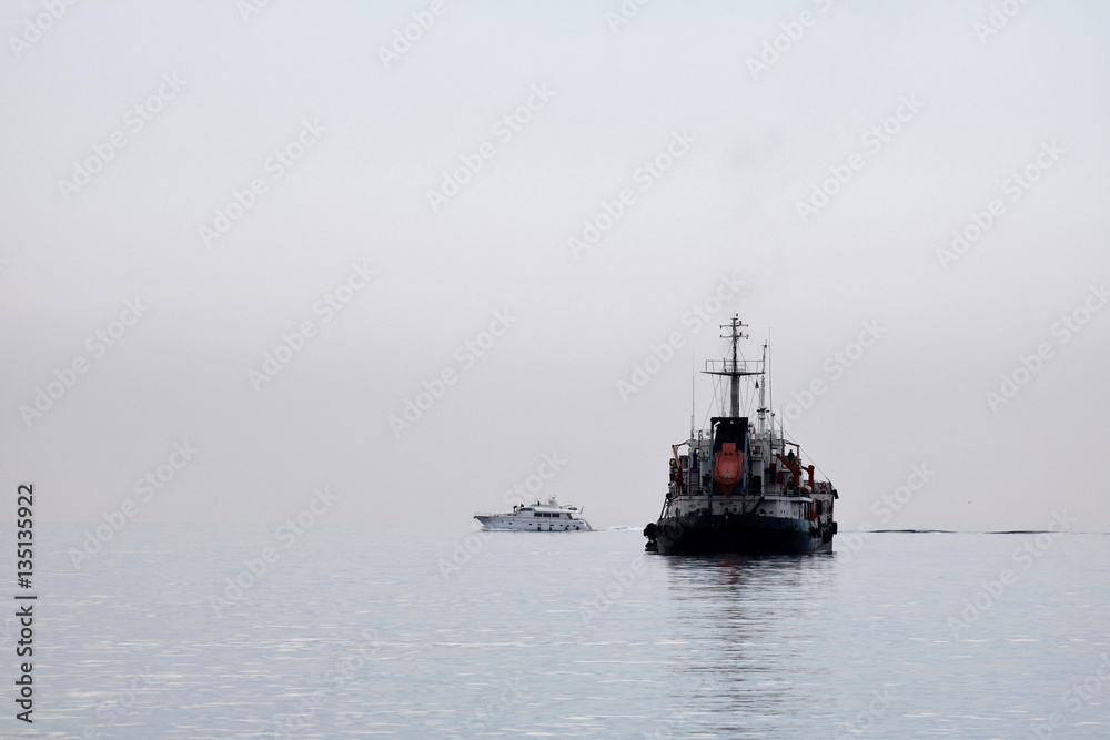 A big ship and small boat crossing paths on calm sea waters on a foggy morning