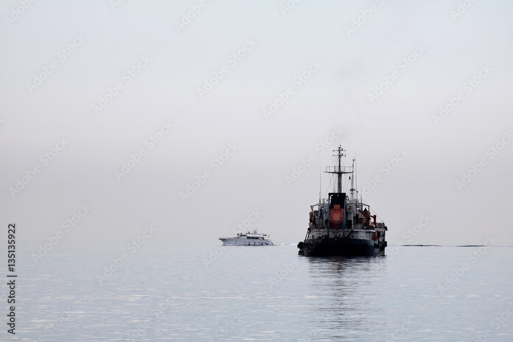 A big ship and small boat crossing paths on calm sea waters on a foggy morning A big ship and small boat crossing paths on calm sea waters on a foggy morning