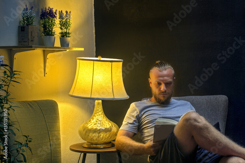 Man reading a book at night. Young man sitting in the living room in the twilight and reading a book. Horizontal indoors shot.  photo