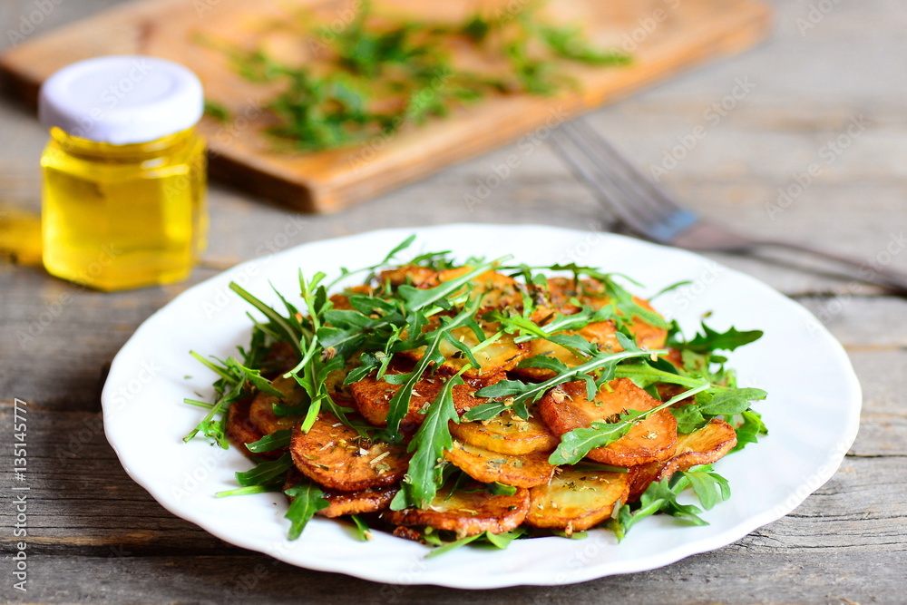 Sliced fried potatoes with arugula and spices on a white plate, a jar of olive oil, fork on wooden table. Homemade food recipe. Cottage fries. Closeup Sliced fried potatoes with arugula and spices on a white plate, a jar of olive oil, fork on wooden table. Homemade food recipe. Cottage fries. Closeup