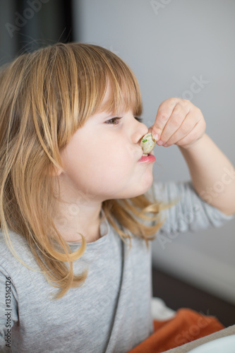 portrait of three years old blonde hungry child face, with grey shirt, eating with her hands and sucking a clam looking 