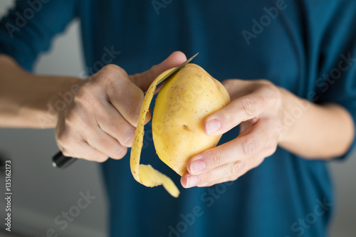detail of hands of woman with blue sweater peeling fresh yellow potato with kitchen knife, horizontal
