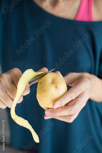 detail of hands of woman with blue sweater peeling fresh yellow potato with kitchen knife, vertical

