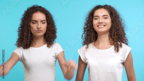 Beautiful Caucasian female twin with curly hair and white t-shirt showing ok with two thumbs while another showing opposite gesture on blue background photo