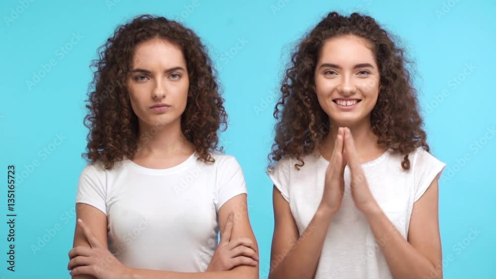 One beautiful Caucasian curly female twin in white t-shirt joyfully claping and showing ok while another one grumpy standing on blue background in slowmotion One beautiful Caucasian curly female twin in white t-shirt joyfully claping and showing ok while another one grumpy standing on blue background in slowmotion