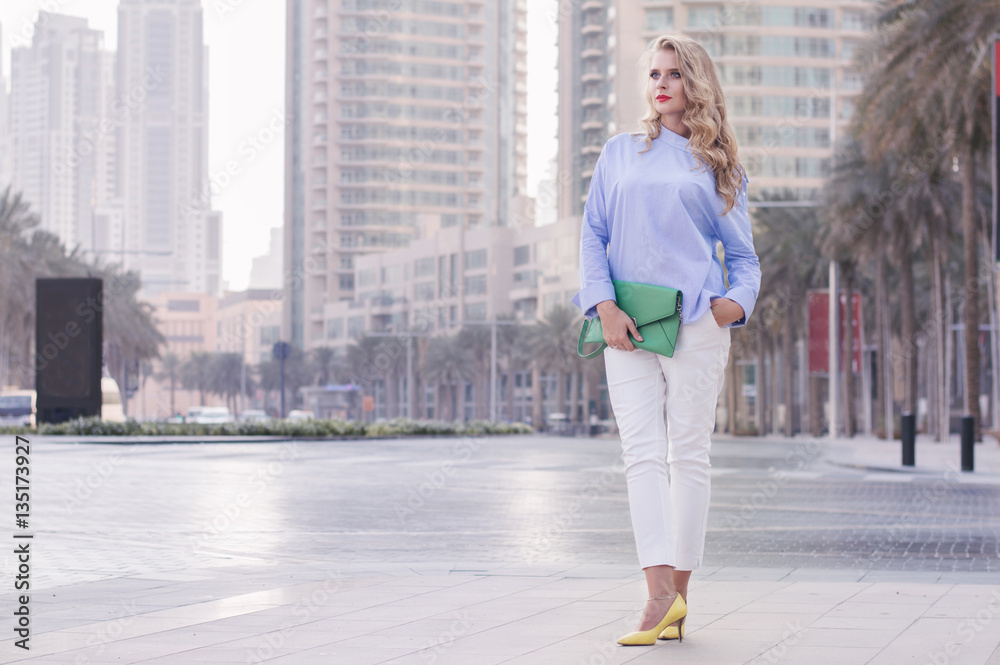 Young european woman with curly blond hair and trendy make up standing near empty road in Dubai downtown, UAE. Stylish business woman in blue blouse, white trousers, yellow shoes and green handbag Young european woman with curly blond hair and trendy make up standing near empty road in Dubai downtown, UAE. Stylish business woman in blue blouse, white trousers, yellow shoes and green handbag