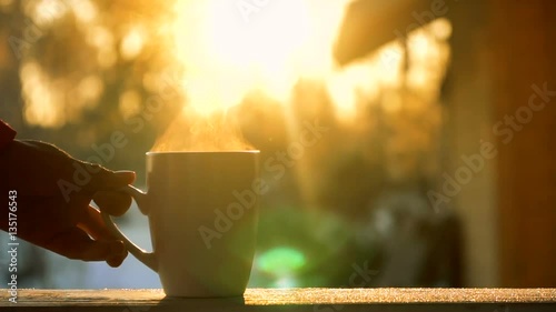 female young hand put white mug of hot drink with steam on wooden desk of stoop of house in the country near forest during winter snowy day under sunlight of sunset