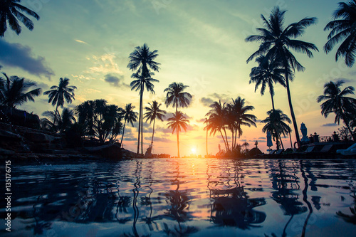 Tropical beach with pool and silhouetted palm trees during sunset. © De Visu